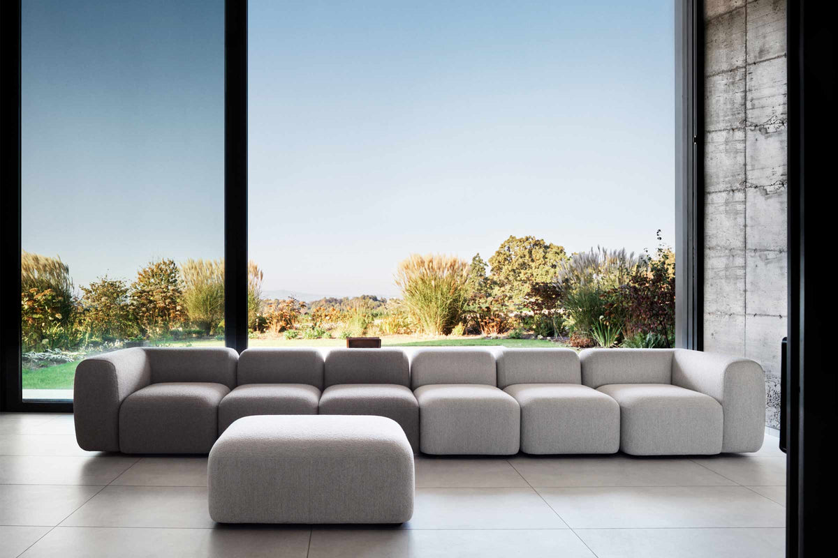 Modular Sofa mit langer Sitzreihe und passendem Hocker vor großer Fensterfront